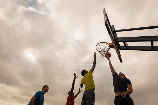 Low angle view of multi-ethnic players playing basketball at basketball court against a cloudy sky — Stock Photo