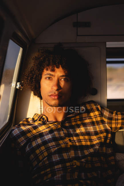 Portrait of a Mixed-race man sitting in camper van and looking at the camera with sun reflects against beach in background on a sunny day — Stock Photo