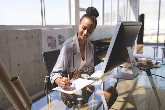 Вид спереду щаслива бізнес-леді змішаної раси, що пише на блокноті, що працює на комп'ютері за столом в офісі — стокове фото