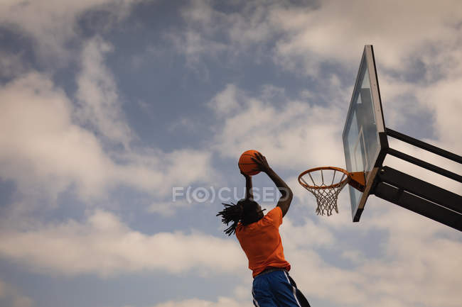 Низкий угол обзора афроамериканского баскетболиста, играющего в баскетбол на баскетбольной площадке во время прыжка, чтобы забить мяч — стоковое фото