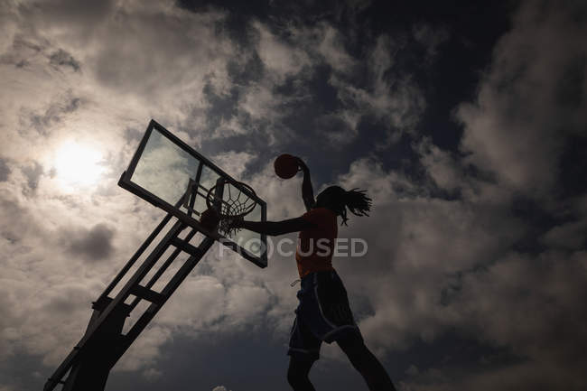 Vista a basso angolo del giocatore di basket afroamericano che gioca a basket sul campo da basket mentre salta per segnare una schiacciata contro il cielo nuvoloso che nasconde il sole — Foto stock