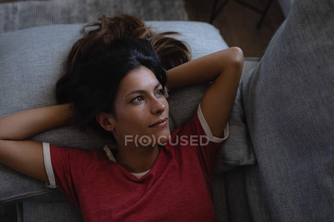 Blick aus der Vogelperspektive auf eine nachdenkliche schöne Mischlingsfrau, die es sich zu Hause auf dem Sofa gemütlich macht — Stockfoto
