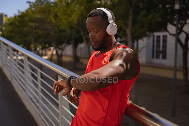 Vista lateral del joven hombre afroamericano en forma con auriculares mirando el reloj inteligente mientras está de pie junto a la barandilla - foto de stock