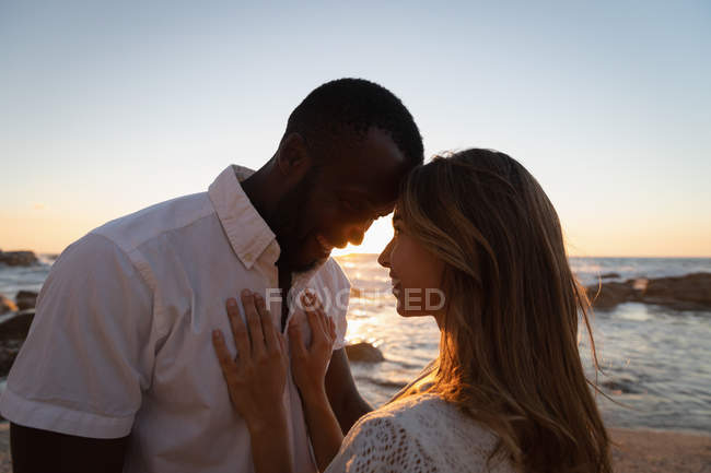 Vista lateral de la pareja multiétnica sonriendo y avergonzándose mutuamente en la playa al atardecer - foto de stock