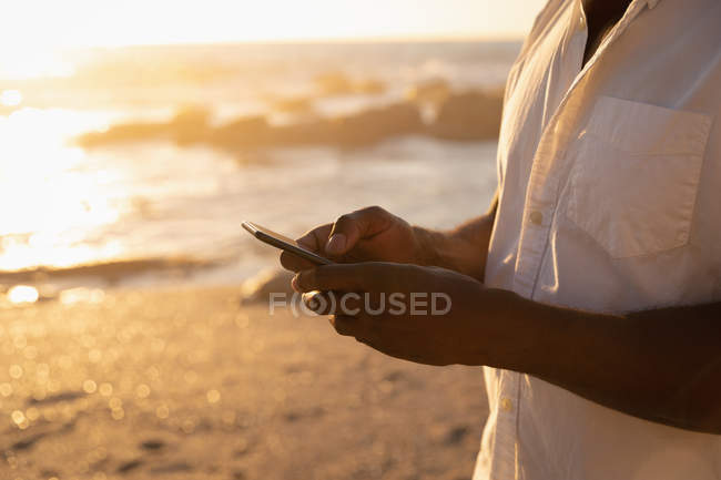 Parte média do homem usando telefone celular na praia ao pôr do sol — Fotografia de Stock