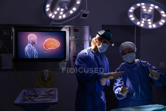 Vista frontal de los cirujanos hablando entre sí durante la cirugía en quirófano en el hospital contra las luces y una pantalla digital - foto de stock