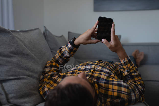 Rückansicht eines jungen kaukasischen Mannes, der sein Handy benutzt, während er zu Hause auf dem Sofa liegt — Stockfoto