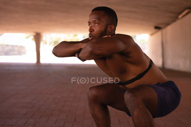 Seitenansicht eines jungen afrikanisch-amerikanischen fitten Mannes beim Kniebeugen unter der Brücke — Stockfoto