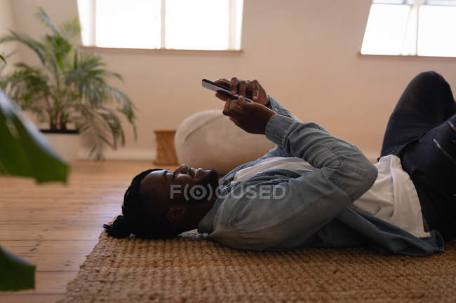 Vista lateral do homem afro-americano bonito usando telefone celular enquanto deitado no chão em casa — Fotografia de Stock