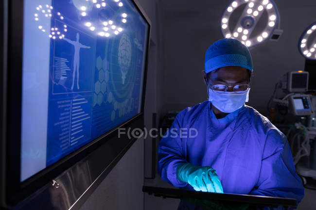 Вигляд спереду чоловічого хірурга, використовуючи цифровий планшетний в операційній в лікарні з великий цифровою екран за ним — стокове фото