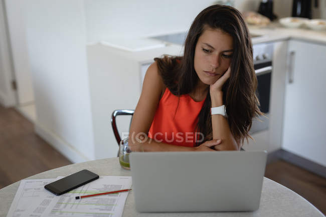 Vista frontal de la triste mujer de raza mixta sentado con el ordenador portátil en la sala de cocina. Ella está pensando - foto de stock
