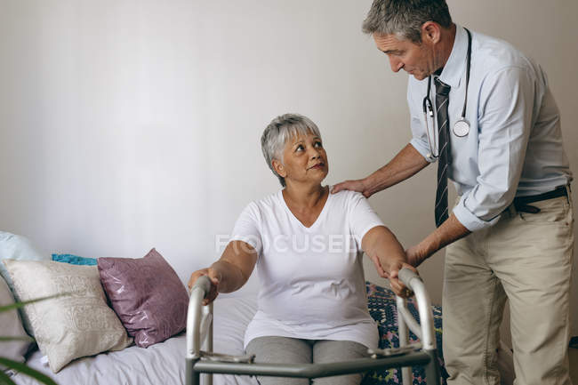 Vista frontale del medico caucasico maschile che aiuta la paziente anziana di razza mista a camminare con walker nella casa di riposo — Foto stock