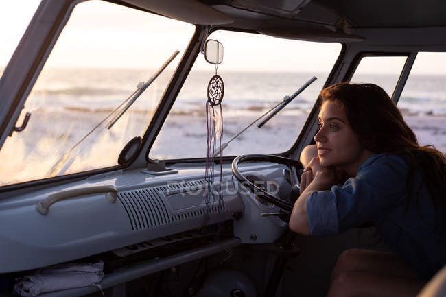 Side view of a thoughtful Caucasian beautiful woman sitting in camper van and leaning on hand wheel against beach in background with sundown — Stock Photo