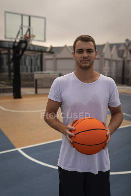 Retrato do jovem caucasiano em pé na frente da câmera enquanto ele está segurando um basquete no playground — Fotografia de Stock