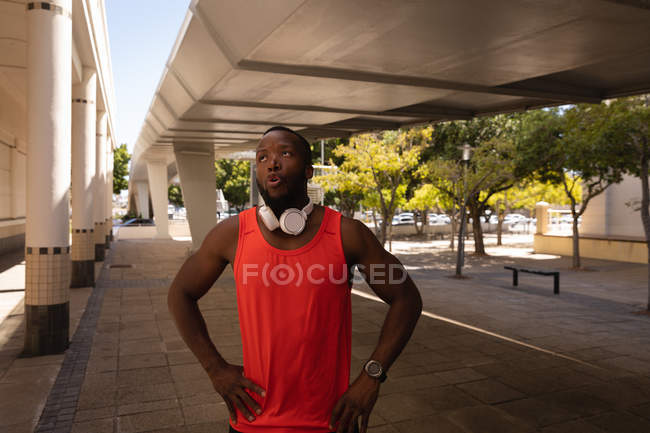 Vista frontale del giovane uomo afro-americano in forma con auricolare in piedi sul marciapiede sotto il ponte — Foto stock