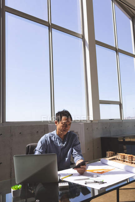 Вид спереди на азиатского архитектора, сидящего за рабочим столом и пользующегося мобильным телефоном в современном офисе. Линейка оранжевого треугольника, геометрический компас, карандаш, ноутбук и архитектурная модель отображаются на столе в современном офисе . — стоковое фото