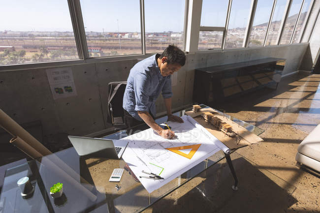 Aus der Vogelperspektive eines männlichen asiatischen Architekten, der am Schreibtisch steht und in einem modernen Büro an einem Entwurf arbeitet — Stockfoto