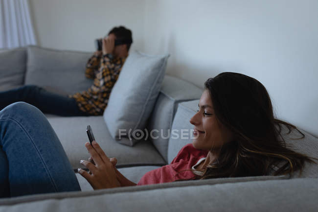 Seitenansicht einer Frau mit gemischter Rasse, die ihr Mobiltelefon benutzt, während ein kaukasischer Mann Virtual-Reality-Headset zu Hause benutzt — Stockfoto