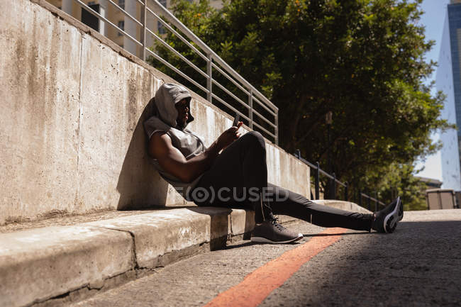 Vista lateral del hombre afroamericano usando el teléfono móvil en el paseo lateral. Él está vestido para hacer footing - foto de stock