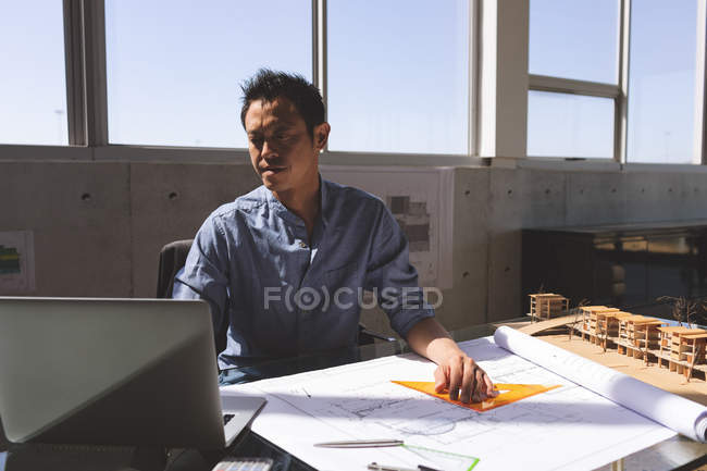 Vista frontal del arquitecto masculino asiático trabajador que utiliza el ordenador portátil mientras trabaja en el plano con regla de triángulo naranja y lápiz en el escritorio en una oficina moderna - foto de stock