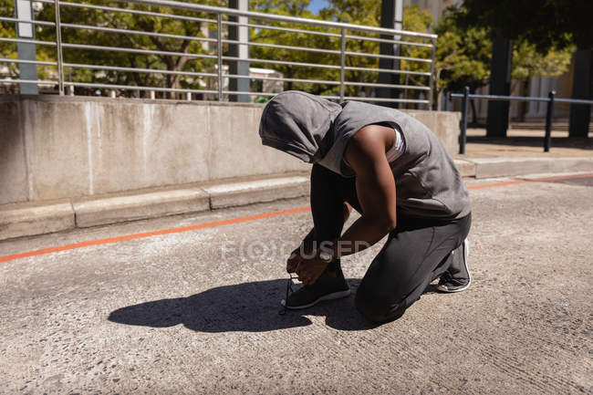 Вид сбоку, как афроамериканец завязывает шнурки на тротуаре. Он одет как бегун. — стоковое фото