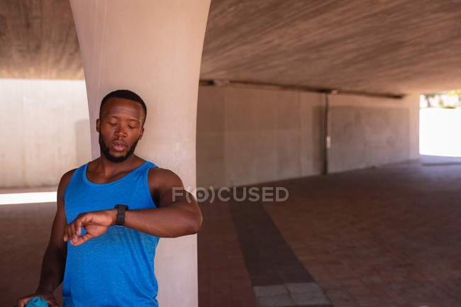 Vista frontale del giovane afro-americano in forma uomo guardando orologio intelligente mentre in piedi sotto il ponte — Foto stock