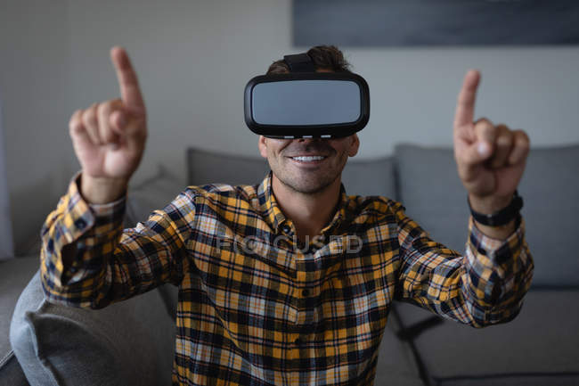 Vista frontale del giovane uomo caucasico utilizzando cuffie realtà virtuale seduto sul divano a casa — Foto stock