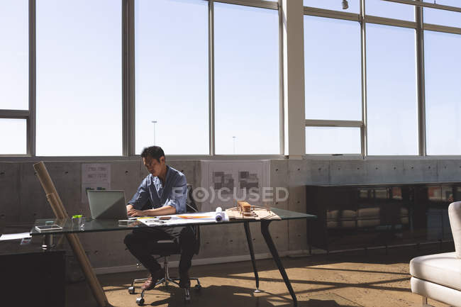 Vista frontal do arquiteto masculino asiático trabalhador sentado na mesa e trabalhando no laptop em um escritório moderno contra o céu azul no fundo — Fotografia de Stock
