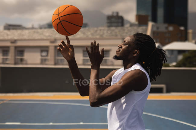 Vista lateral do jogador de basquete afro-americano equilibrando bola no dedo na quadra de basquete contra a cidade embaçada no fundo — Fotografia de Stock