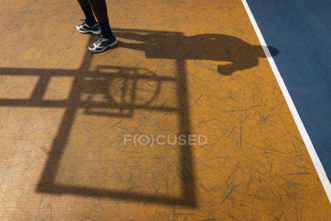 Низька частина баскетболіста стоїть на баскетбольному майданчику проти його тіні, а також баскетбольна тінь, що відображається на дитячому майданчику — стокове фото