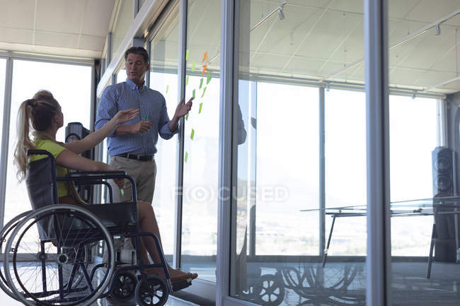 Vista lateral de la mujer rubia caucásica discapacitada y el ejecutivo masculino caucásico discutiendo sobre notas adhesivas en la oficina - foto de stock