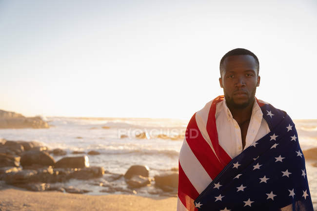O retrato do homem afro-americano envolvia a bandeira americana enquanto estava na praia ao pôr-do-sol. Ele está olhando para a câmera — Fotografia de Stock
