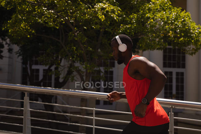Vista lateral de un joven afroamericano en forma con auriculares que corren contra la barandilla en un día soleado - foto de stock