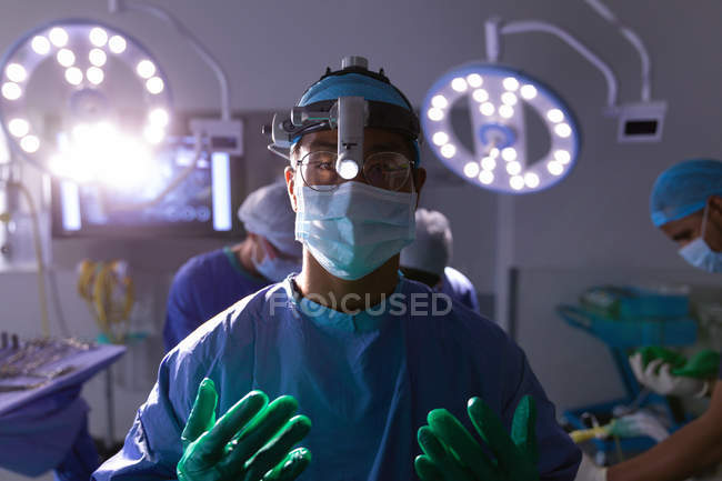 Retrato del cirujano asiático de pie y mirando la cámara en el quirófano en el hospital contra los cirujanos que actúan en segundo plano - foto de stock
