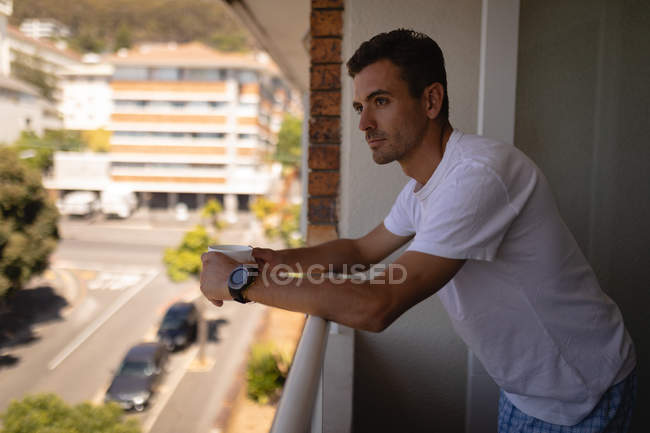 Side view of handsome young Caucasian man with cup of coffee standing in balcony at home. He is looking away — Stock Photo