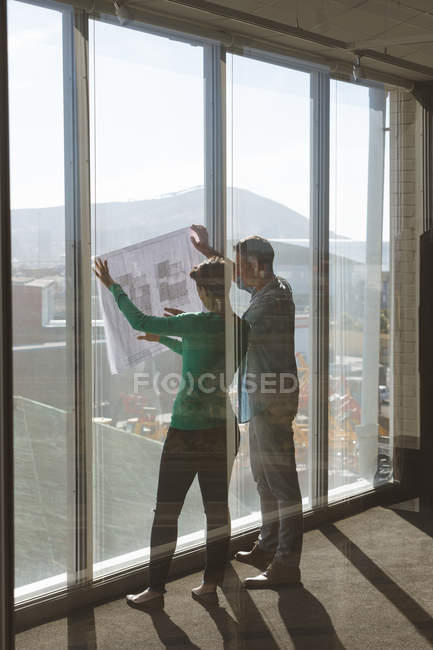 Vista trasera de los arquitectos caucásicos sosteniendo el plano contra una ventana y discutiendo sobre ello en la oficina - foto de stock
