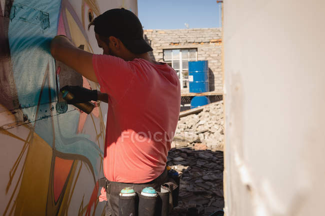 Vista lateral do jovem artista de graffiti caucasiano pintura spray na sala de parede resistida — Fotografia de Stock