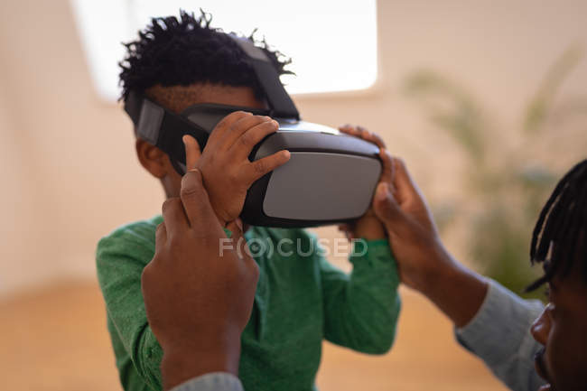 Vista lateral del padre afroamericano ayudando a su hijo a usar auriculares de realidad virtual en casa - foto de stock