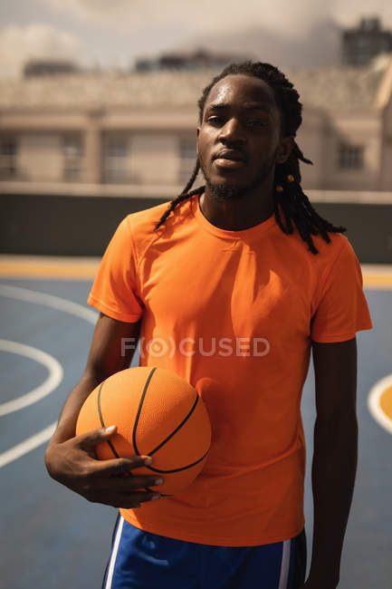 Retrato de jogador de basquete afro-americano de pé com basquete no playground contra a cidade embaçada no fundo. Ele está olhando para a câmera — Fotografia de Stock