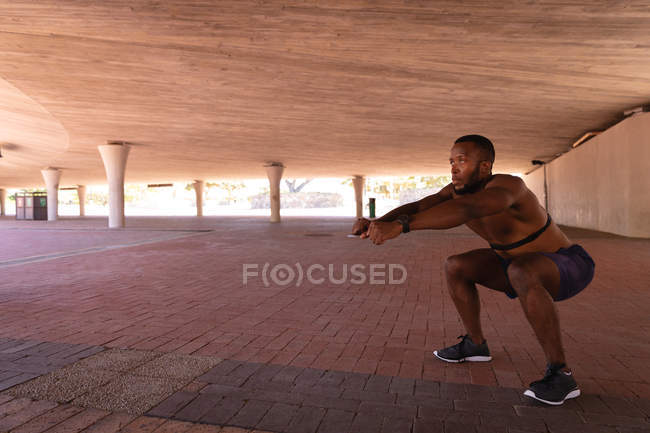 Seitenansicht eines jungen afrikanisch-amerikanischen fitten Mannes beim Kniebeugen unter der Brücke — Stockfoto