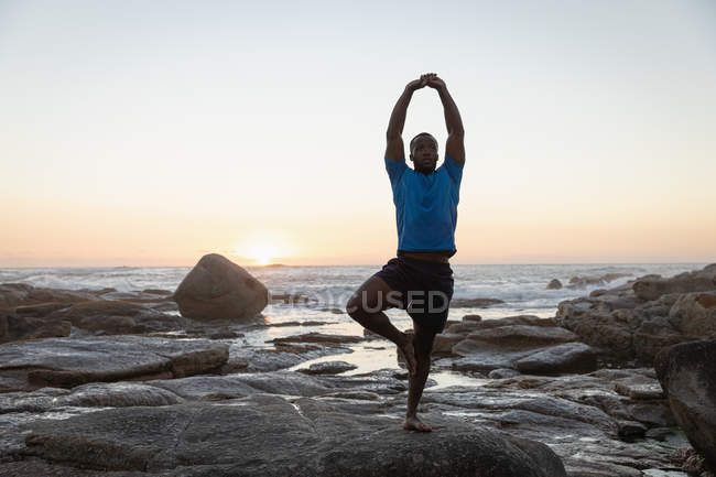 Vista frontale del giovane uomo afro-americano che fa yoga in spiaggia su rocce al tramonto — Foto stock