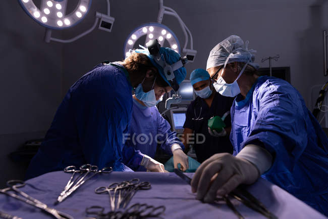 Низкий угол обзора хирургов, сконцентрированных на выполнении операции в операционной в больнице с ножницами на переднем плане — стоковое фото