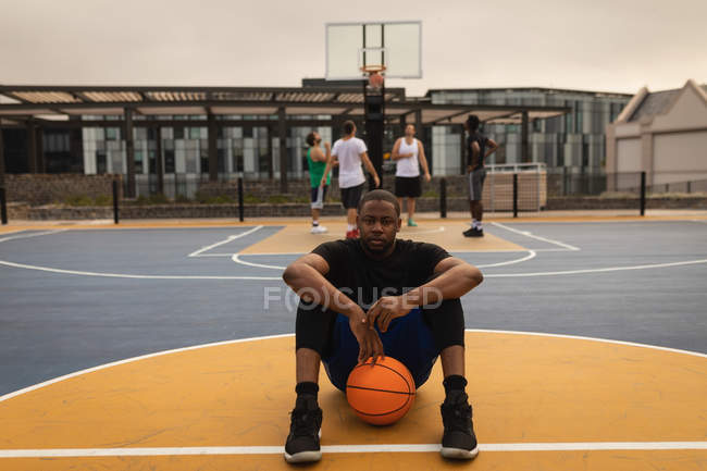 Retrato de un jugador afroamericano confiado en el baloncesto sentado en la cancha de baloncesto con jugadores detrás de él - foto de stock