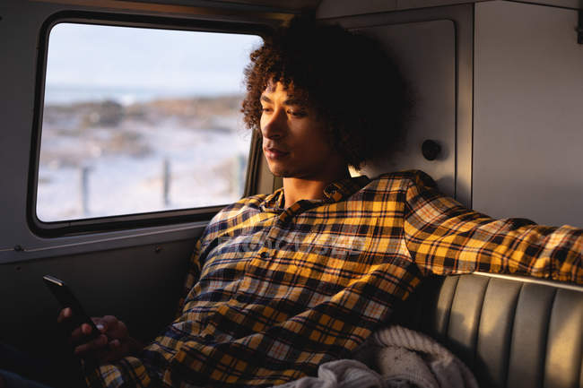Side view of a Mixed-race man using mobile phone in a camper van against beach on a sunny day in background — Stock Photo