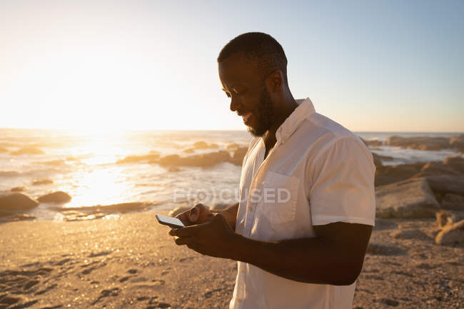 Vista lateral del joven afroamericano usando teléfono móvil en la playa al atardecer. Él sonríe. - foto de stock