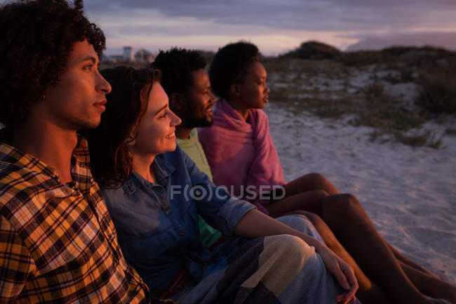 Side view of multi-ethnic friends group sitting on beach while they are looking away ocean with sun reflecting on their face — Stock Photo