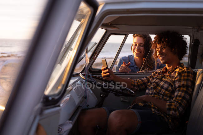 Vista lateral de un hombre de raza mixta en una caravana tomando una selfie con una mujer caucásica apoyada fuera de la ventana contra la playa en el fondo - foto de stock