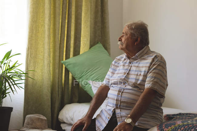 Vista lateral de um homem caucasiano sênior olhando para fora da janela enquanto sentado sozinho na cama do lar de idosos — Fotografia de Stock