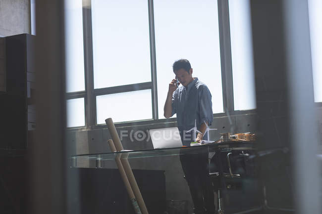 Vista frontal del arquitecto asiático de pie y hablando por teléfono móvil mientras trabaja en el escritorio en una oficina moderna - foto de stock