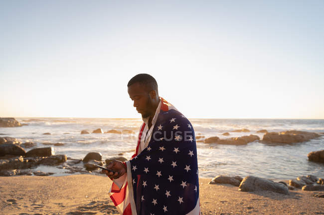 Seitenansicht eines jungen afrikanisch-amerikanischen Mannes, der die amerikanische Flagge eingehüllt hat, während er sein Handy am Strand bei Sonnenuntergang benutzt — Stockfoto